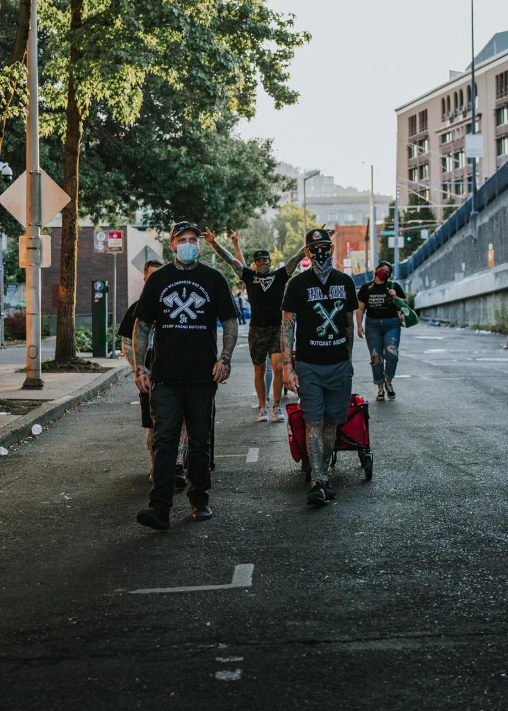 Six people walk along a street together in casual clothing such as t-shirts and shorts or jeans. One pulls a red cart behind them.